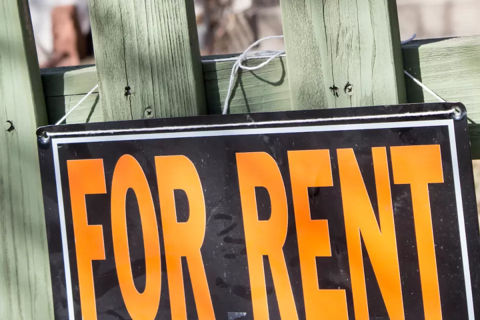Close-up of orange and black For Rent sign on green wood fence.